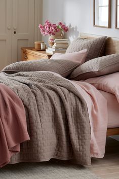 a bed with pink sheets and pillows in a bedroom next to a dresser filled with flowers