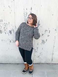 a woman standing in front of a white wall wearing black and grey sweater, leggings, and brown boots