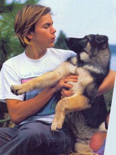 a man holding a dog in his arms while sitting on the ground next to trees