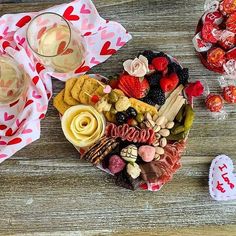 a heart shaped platter filled with assorted snacks and wine glasses on a wooden table