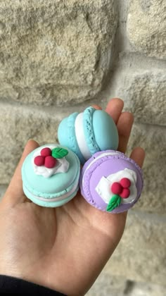 a hand holding three decorated macaroons in front of a stone wall