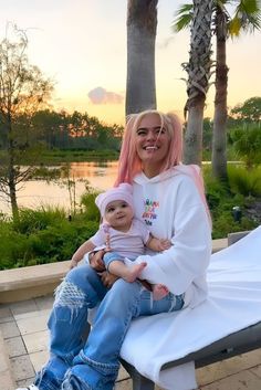 a woman with pink hair sitting on a bench holding a baby and smiling at the camera