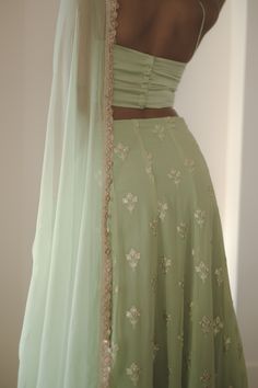 I think we just found your new mehendi set! Ira is an enchanting sage green 3-piece gotta patti lehenga set. The ensemble comes with an intricately embroidered skirt, a chic cowl neck blouse, and a matching chiffon dupatta with a gotta patti border. Ira embodies timeless elegance and contemporary allure. Blouse, lehenga, and dupatta included. Light Green Indian Outfit, Halter Top Lehenga, Aesthetic Chaniya Choli, Green Chikankari Embroidery Lehenga For Reception, Aesthetic Lehenga Designs, Simple Dresses Indian, Simple Lengha Design, Onam Dress Ideas, Light Green Lehenga