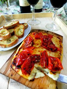 a wooden cutting board topped with pizza and bread next to two glasses of red wine