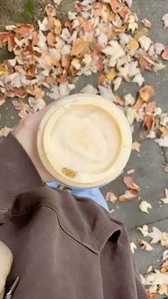 a person holding a cup of coffee in their hand with leaves on the ground behind them