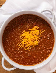 a white bowl filled with chili and cheese on top of a wooden table next to a napkin