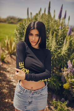 a beautiful young woman standing in front of purple flowers wearing a black shirt and jean shorts