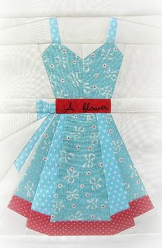 a dress made out of paper with red ribbon and blue flowers on the front, sitting on top of a piece of white fabric