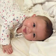 a smiling baby laying on top of a bed covered in white sheeted sheets and blankets