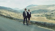 two business people walking down the road in front of some hills and valleys, one man is wearing a suit