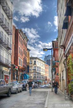 the people are walking down the narrow street in the city with cars parked on both sides