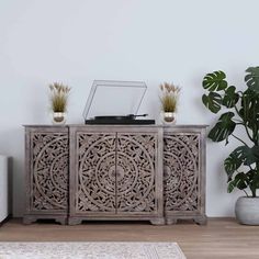 a wooden sideboard with carvings on it in front of a white wall and potted plants