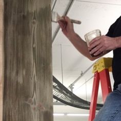 a man sitting on a step ladder painting the side of a wooden structure with white paint