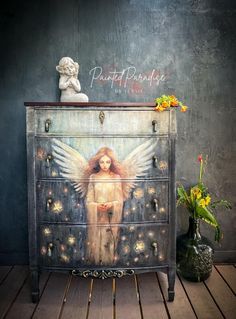 an angel painted on the side of a dresser next to a vase with flowers in it