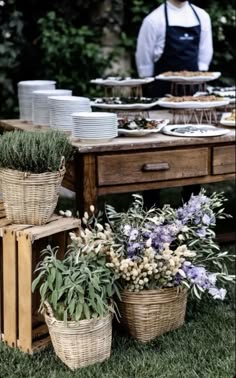 a table topped with lots of baskets filled with flowers next to a man in an apron