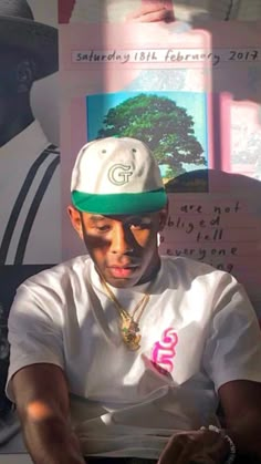 a man wearing a white shirt and green hat sitting in front of a wall with pictures on it