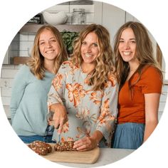 three women are standing together in the kitchen and one is cutting some food on a board