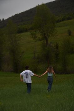 two people holding hands walking through a field with trees and hills in the back ground