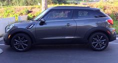 a small gray car parked in a parking lot next to a white fence and trees