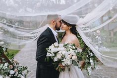 a bride and groom kissing under the veils of their wedding arch with white flowers