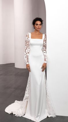 a woman standing in front of a white wall wearing a long sleeved wedding dress