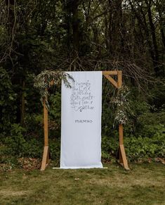 a white sign with writing on it sitting in the grass next to some bushes and trees