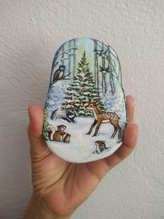 a hand holding up a painted rock with animals around the christmas tree on it's side