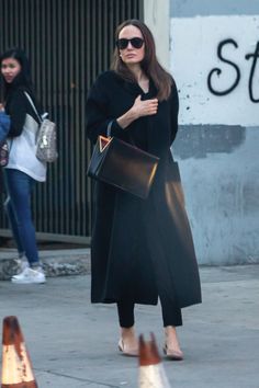 a woman in black coat and sunglasses walking down the street with her hand on her purse