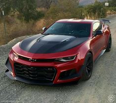 a red chevrolet camaro is parked on the side of a dirt road with trees in the background