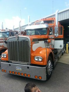 an orange semi truck parked in front of a building with people looking at the trucks