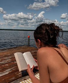 a woman sitting on a dock reading a book next to the water with clouds in the sky