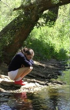 a woman crouches down to look at the water while standing in front of a tree