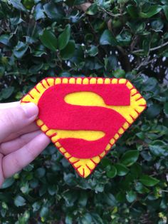 a hand holding a small felt superman symbol in front of green bushes and shrubbery