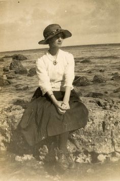 an old black and white photo of a woman sitting on rocks near the ocean with her legs crossed