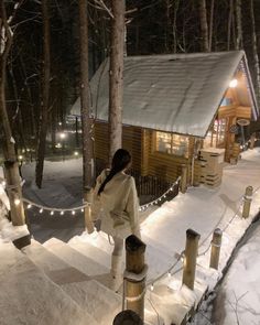 a woman is standing in the snow near a log cabin at night with lights on