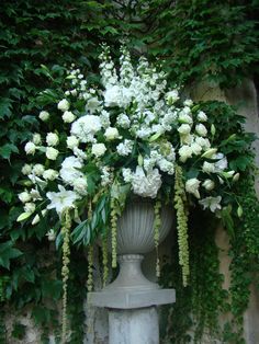 white flowers and greenery are in a vase on a stone pedestal next to an ivy covered wall