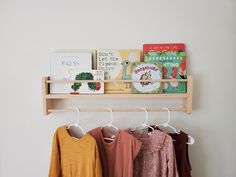 three children's shirts hanging on a wooden shelf next to books and other items