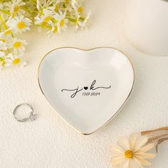 a white heart shaped dish with the word joy on it next to daisies and a wedding ring