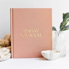 a pink travel journal sitting on top of a table next to a vase with a plant in it