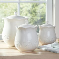 three white vases sitting on top of a table next to a book and window