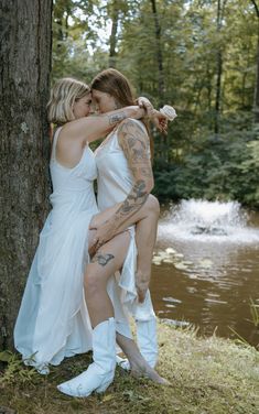 two women in white dresses hugging each other near a tree and water with trees behind them