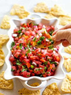 a hand holding a tortilla chip over a white bowl filled with salsa and chips