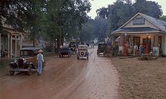 old cars parked on the side of a dirt road in front of small buildings and trees