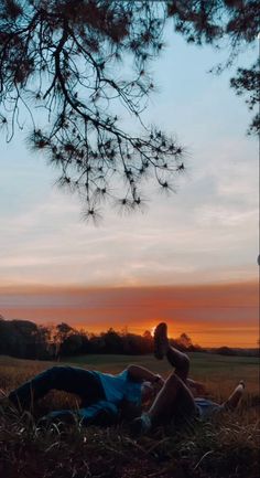 two people are laying in the grass under a tree at sunset, with one person holding an object up to his head