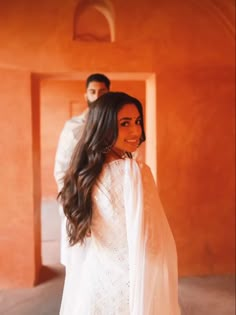 a man standing next to a woman in a white dress with long hair and an orange wall