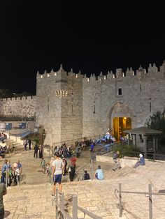 people are walking around in front of an old castle at night with the lights on