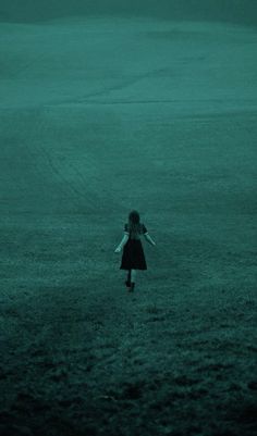 a girl in a black dress is walking through the dark grass with her arms outstretched