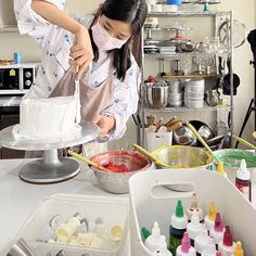 two women in aprons and masks are decorating a white cake with icing