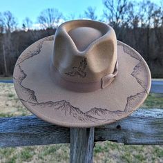 This wide brim fedora makes a great addition to any nature lover's wardrobe. The neutral tan material burns a gorgeous shade of dark brown allowing the design to stand out. Each mountain is intricately burned by hand with a pyrography machine using various tips to achieve depth and rock-like texture. The outline of an eagle flying is burnt on the front of the crown. The design is original and completely free-handed. For a beautiful finishing touch the hat is adorned with a  tan bow hatband.  Hat Materials: polyester Hat has a 58-60cm crown, but fits most with an adjustable tie band inside Hat Brim: Roughly 4 inches Colors may vary slightly from screen to screen Burnt Cowboy Hat, Felt Hat Burning Designs, Felt Hat Burning, Hat Burning Designs, Burned Hats, Hat Burning, Bird Watcher Gifts, Eagle Flying, Nature Gifts