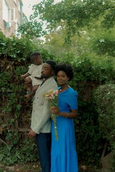 a man and woman holding a baby standing next to each other in front of some bushes